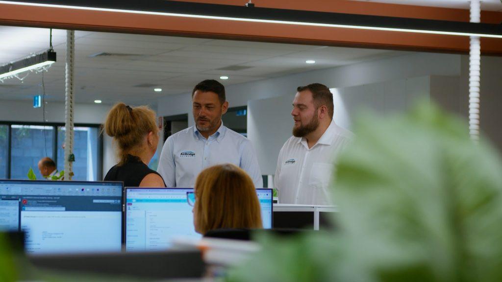 Doug and Stephen standing in a modern office, engaging in a conversation with a woman on the screens in front of them, as they discuss details with a second woman in person.